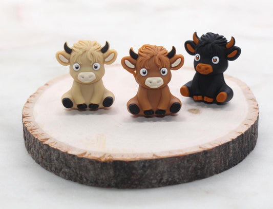 May include: Three small toy figurines of cows. One cow is brown, one is black, and one is light brown. The cows are all standing on a wooden surface.