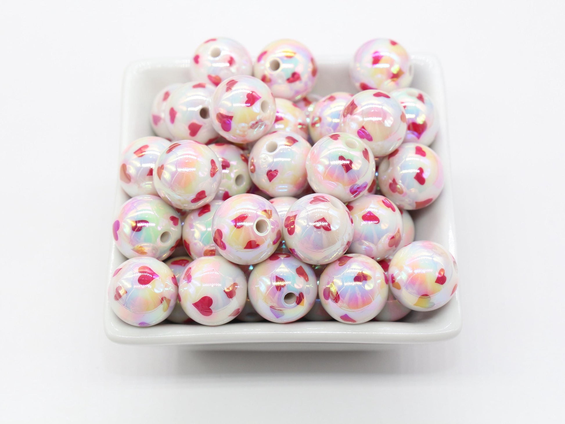 A collection of multicolored beads with heart patterns in a white square dish on a white background.