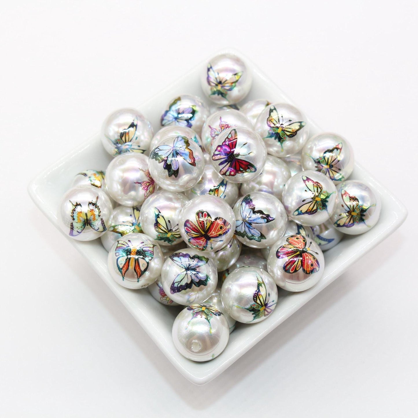 A group of white marbles with colorful butterfly patterns in a square dish on a white background.