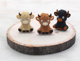 May include: Three small toy figurines of cows. One cow is brown, one is black, and one is light brown. The cows are all standing on a wooden surface.