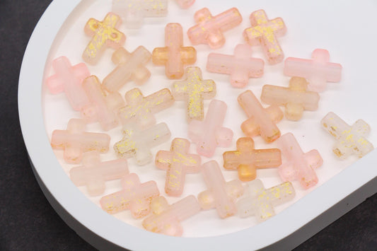 May include: A collection of translucent cross-shaped beads in shades of pink, peach, and white, with gold glitter accents. The beads are scattered on a white, rounded tray, creating a delicate and decorative display. Ideal for jewelry making or craft projects.