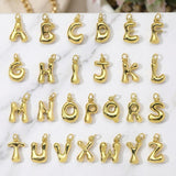 May include: A collection of twenty-six individual gold-toned metal letter charms, each featuring a rounded, puffed, and glossy finish. Every charm includes a small circular jump ring at the top for attachment, displaying the full alphabet from A to Z.